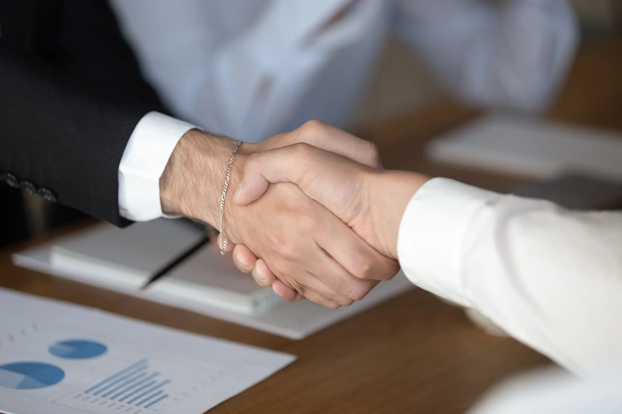 Two business professionals shaking hands over documents with charts, symbolizing partnership, agreement, and successful business collaboration.